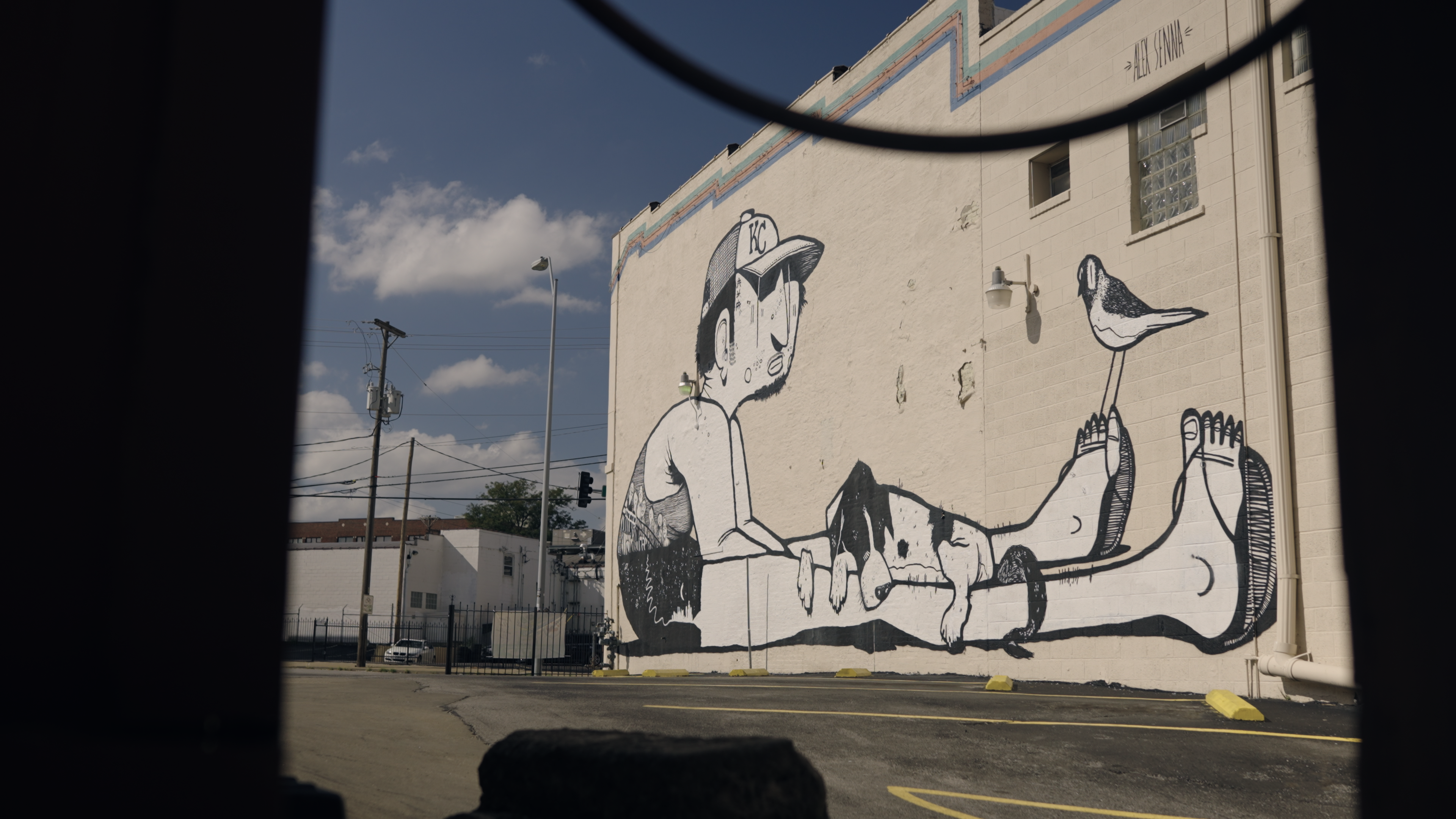 A large mural on an exterior brick wall of a figure sitting with his legs outstretched and dog sitting in his lap