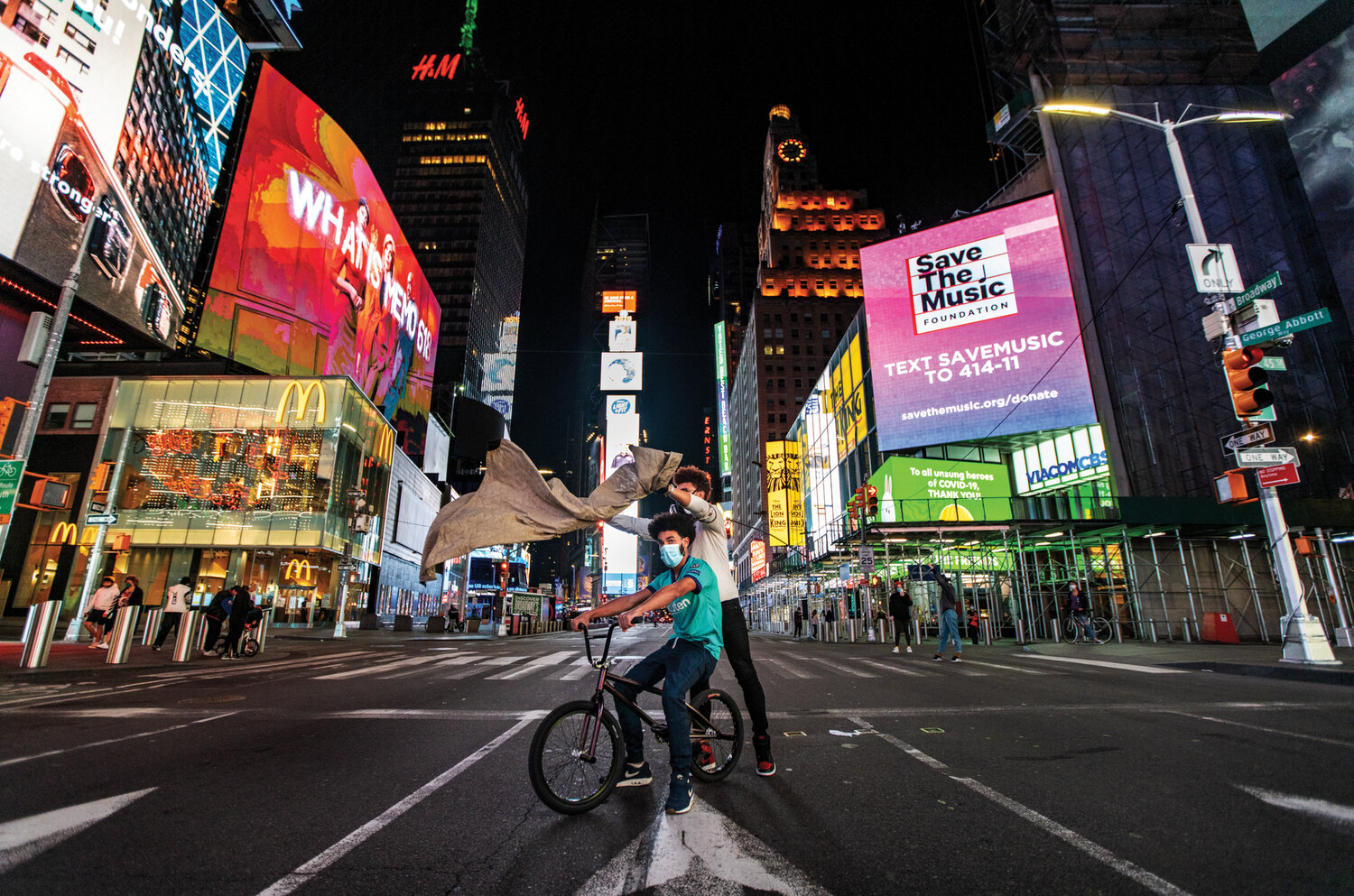 Julien Howard a.k.a. Velo Barber placing a barber cape over a person sitting on a bicycle in the center of NYC, in the center of Times Square, cross roads of the world