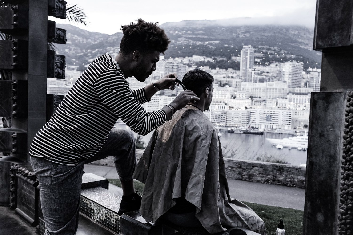 Julien Howard a.k.a. Velo Barber wearing a striped shirt and using shears to cut a persons hair in front of a terrace opening with a beautiful view of a city scape on a mountain in the background